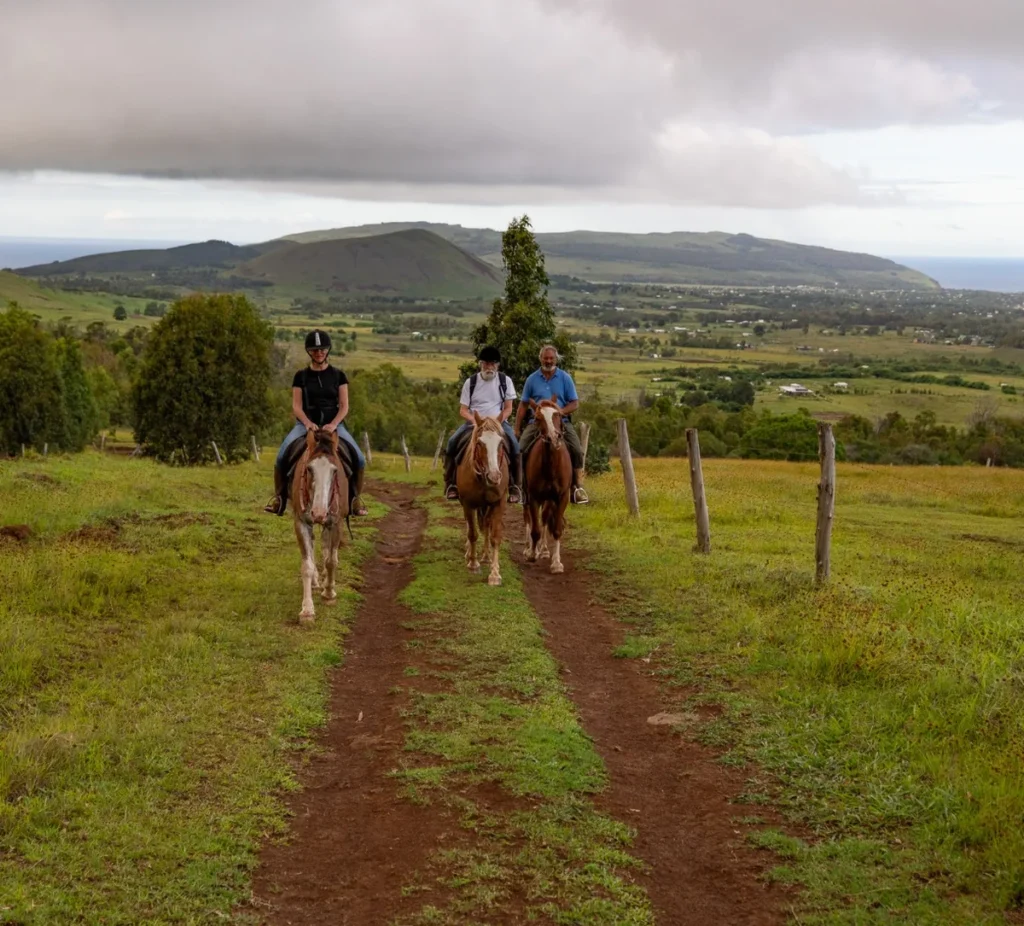 Tres jinetes bajando por un camino de tierra, con vista sobre Hanga Roa, los conos volcánicos y el océano.