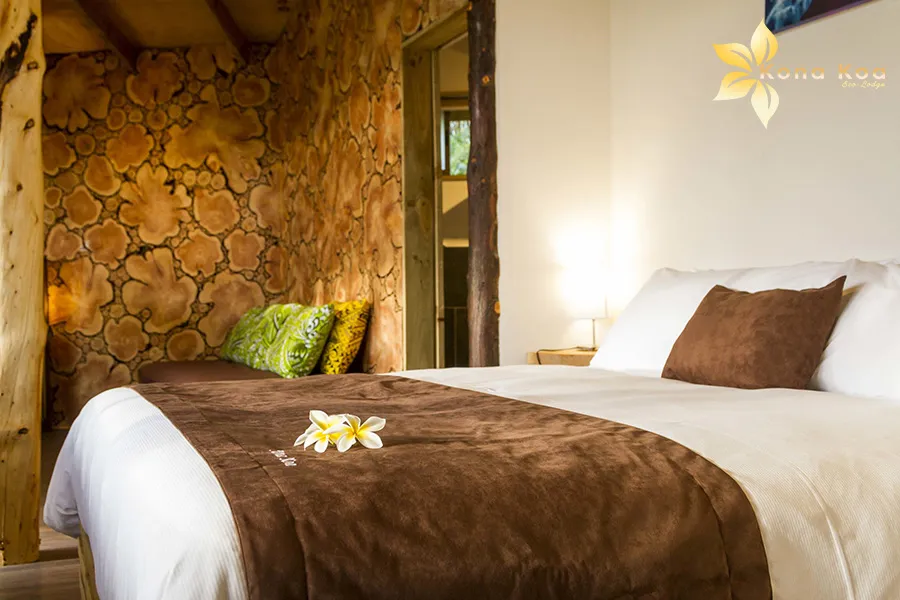 Double bed in a bungalow at Kona Koa Eco Lodge, log wall and a frangipani flower on the sheets.