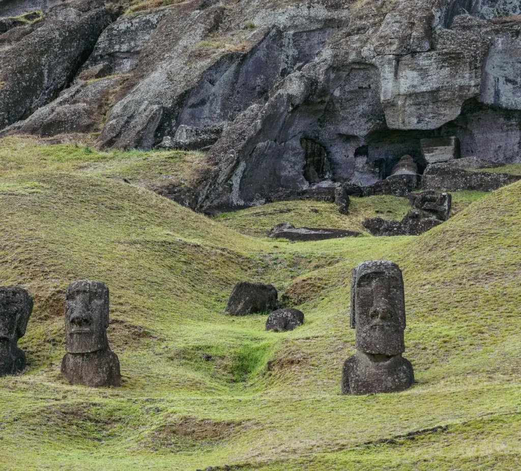 Rock face hollowed with cavities at Rano Raraku, three moai heads emerging from the green grass in the foreground.