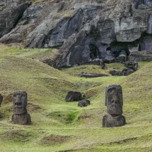 Rock face hollowed with cavities at Rano Raraku, three moai heads emerging from the green grass in the foreground.